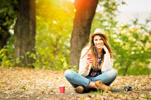 Stylish young woman sitting in the woods using cell phone