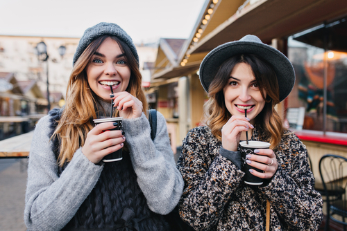 Two young women smiling and sipping drinks outside